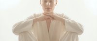 Young adult male with fair complexion wearing a cream linen shirt, hands positioned at chest in a contemplative gesture against neutral background.