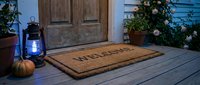 Cozy front porch with a welcome doormat, glowing lantern, small pumpkin, and blooming roses by a wooden door in warm autumn light.
