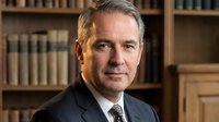 Middle-aged Caucasian male professional in dark business suit and tie, positioned against wooden bookshelves in law office or corporate library setting, suitable for business portraits and professional headshots.