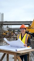 Female construction engineer in safety gear reviewing blueprints at building site with heavy machinery and concrete structures in background.