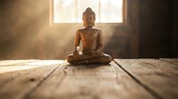 Buddha statue in meditation pose on wooden floor with warm sunlight streaming through window creating peaceful spiritual atmosphere for mindfulness practice.