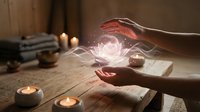 Hands holding glowing magical energy orb with floating lotus flower in cozy candlelit room with books creating mystical spiritual atmosphere.