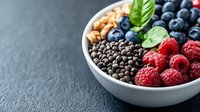Fresh raspberries, blueberries, chocolate chips and granola in white ceramic bowl on dark stone background with basil leaves, healthy breakfast concept.