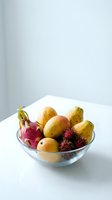 Glass bowl filled with fresh tropical fruits including mangoes, pears, dragon fruit and rambutan on white surface. Healthy eating and nutrition concept.