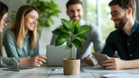 Young diverse business team having casual meeting at office desk with small green plant in focus, discussing sustainable development and eco initiatives.