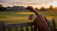 Vintage leather golf bag with clubs resting against wooden fence at sunset overlooking green course and clubhouse for sports and recreation themes.