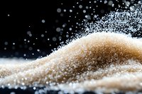 Brown sugar crystals scattered on dark background with flying particles creating dynamic composition, macro food photography for culinary and sweet concepts.