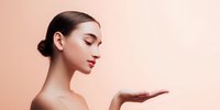 Young Asian woman with sleek hair bun and bare shoulders presenting with open palm against peach background for beauty product promotion.