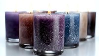 Glittering scented candles in glass holders with burning wicks arranged in row, featuring purple, brown, and blue colors on white surface. Soft focus background.