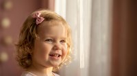 Happy blonde toddler girl with pink bow smiling joyfully by window in warm natural light for family lifestyle photography and childhood memories.