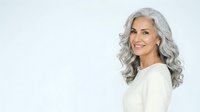 Mature woman with long gray wavy hair, fair complexion, wearing cream-colored top, smiling at camera against white background for beauty, wellness, and lifestyle marketing.