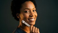 Young African American woman applying makeup with brush, smiling confidently against dark background. Professional beauty routine and cosmetic application concept.