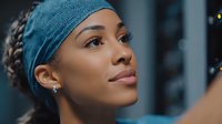 Young mixed-race woman in teal headband taking selfie with smartphone, natural makeup and earrings, urban evening background with bokeh lights.
