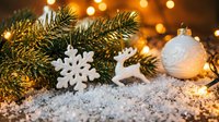 Festive holiday arrangement featuring evergreen branches, white snowflake and reindeer ornaments, pearl bauble, and warm golden lights on snowy surface. Perfect for seasonal marketing and holiday designs.