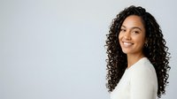 Young woman with long curly dark hair and warm smile wearing white ribbed sweater, isolated on light background for portrait and lifestyle use.
