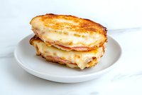 Grilled ham and cheese sandwich with melted cheese oozing out between toasted bread slices, served on white plate against marble background. Perfect for menu design.