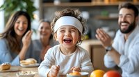 Joyful child wearing white headband laughs while family enjoys breakfast together at kitchen table with pastries and fresh fruit in morning light.