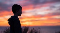 Young boy silhouette against dramatic sunset sky with orange and pink clouds. Child profile contemplating nature during golden hour twilight moment.