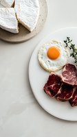 Fresh breakfast composition with sunny side up egg, raw marbled beef steak cuts, and soft brie cheese wheel on ceramic plates against white background.