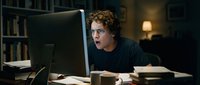 Young man with curly hair and glasses, appearing surprised or shocked while working at desktop computer in home office with bookshelves and warm lighting.