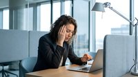 Stressed businesswoman with headache working on laptop in modern office. Professional burnout and workplace pressure concept for corporate wellness campaigns.