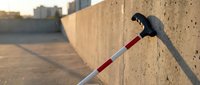 Visually impaired person using white and red mobility cane walking along concrete wall in urban environment during golden hour lighting.