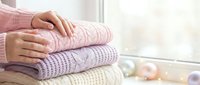 Woman hands folding stack of cozy knit sweaters in soft pastel colors on white surface near window. Seasonal wardrobe organization and home tidying concept.