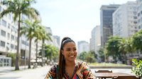 Young woman with dark braided hair and medium brown skin tone wearing a colorful patterned shirt smiles while standing on an urban street lined with palm trees and modern apartment buildings during daytime.