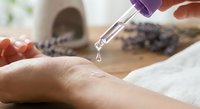 Hands applying lavender essential oil drops from purple dropper bottle onto skin for aromatherapy massage and natural wellness treatment.