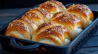 Fresh baked sesame bread rolls in blue baking pan with golden brown crust and shiny surface, homemade soft yeast dough pastry on dark background.