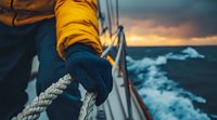 Sailor wearing protective gloves grips rope on sailing yacht during stormy weather at sunset, dramatic ocean waves and moody sky in background.