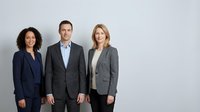 Professional business team of three diverse colleagues in formal suits standing confidently against neutral background for corporate presentations.