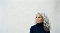 Mature woman with long silver wavy hair and fair complexion wearing dark turtleneck against neutral background, suitable for lifestyle, beauty, and wellness content.