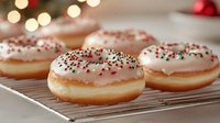 Fresh baked donuts with pink frosting and festive sprinkles on cooling rack against blurred holiday lights background create cozy winter atmosphere.