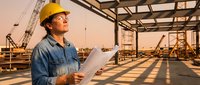 Construction worker in yellow hard hat reviewing blueprints at building site during golden hour. Professional contractor planning project development.