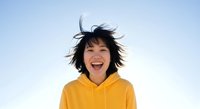 Young woman with East Asian appearance wearing bright yellow hoodie, laughing with mouth open and windblown black hair against clear blue sky background, expressing joy and happiness.