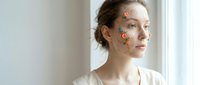 Young Caucasian woman with orange floral face stickers looking thoughtfully out window in bright modern interior for skincare and beauty concepts.