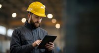 Industrial worker in yellow hard hat using digital tablet in manufacturing facility with bokeh lights background for construction technology applications.