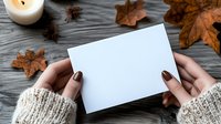 Woman hands holding blank white card on rustic wooden table with autumn leaves, candle and star anise. Cozy fall mockup for greeting cards, invitations.