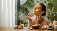 Young Asian woman applying facial mask with brush in spa setting surrounded by tropical plants and candles for natural skincare routine.