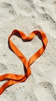Orange satin ribbon forming heart shape on sandy beach surface for romantic Valentine's Day wedding anniversary love celebration greeting cards.