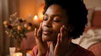 Young African American woman applying skincare cream in cozy bedroom with warm lighting and candles creating relaxing spa atmosphere for evening routine.