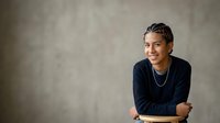 Young man with dark hair and South Asian ethnicity wearing navy blue sweater with chain necklace, smiling at camera against neutral gray background for portrait and professional use.