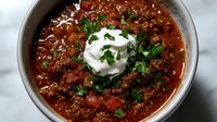 Hearty bowl of homemade chili con carne topped with sour cream and fresh chopped parsley, served on marble counter. Rich meaty texture with beans in tomato sauce.