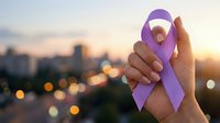 Hand holding purple awareness ribbon against blurred cityscape at sunset, symbolizing support for health causes and charitable campaigns.