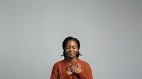 Young woman with dark braided hair and medium brown skin tone wearing rust-colored sweater, hand on chest, smiling warmly against neutral gray background for portrait and lifestyle use.