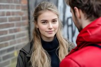 Young woman with blonde hair wearing black turtleneck and jacket smiles warmly while engaged in conversation with man in red coat against brick wall background.