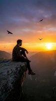 Young man sitting on mountain cliff edge watching sunset with birds flying overhead in dramatic sky for adventure and freedom concepts.