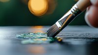 Paintbrush with black handle dipping into teal and gold paint on dark surface, soft bokeh lights in background create artistic atmosphere for creative projects.