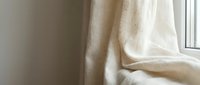 Soft natural linen curtain fabric draping elegantly by bright window creating warm minimalist home interior atmosphere with gentle sunlight filtering through.